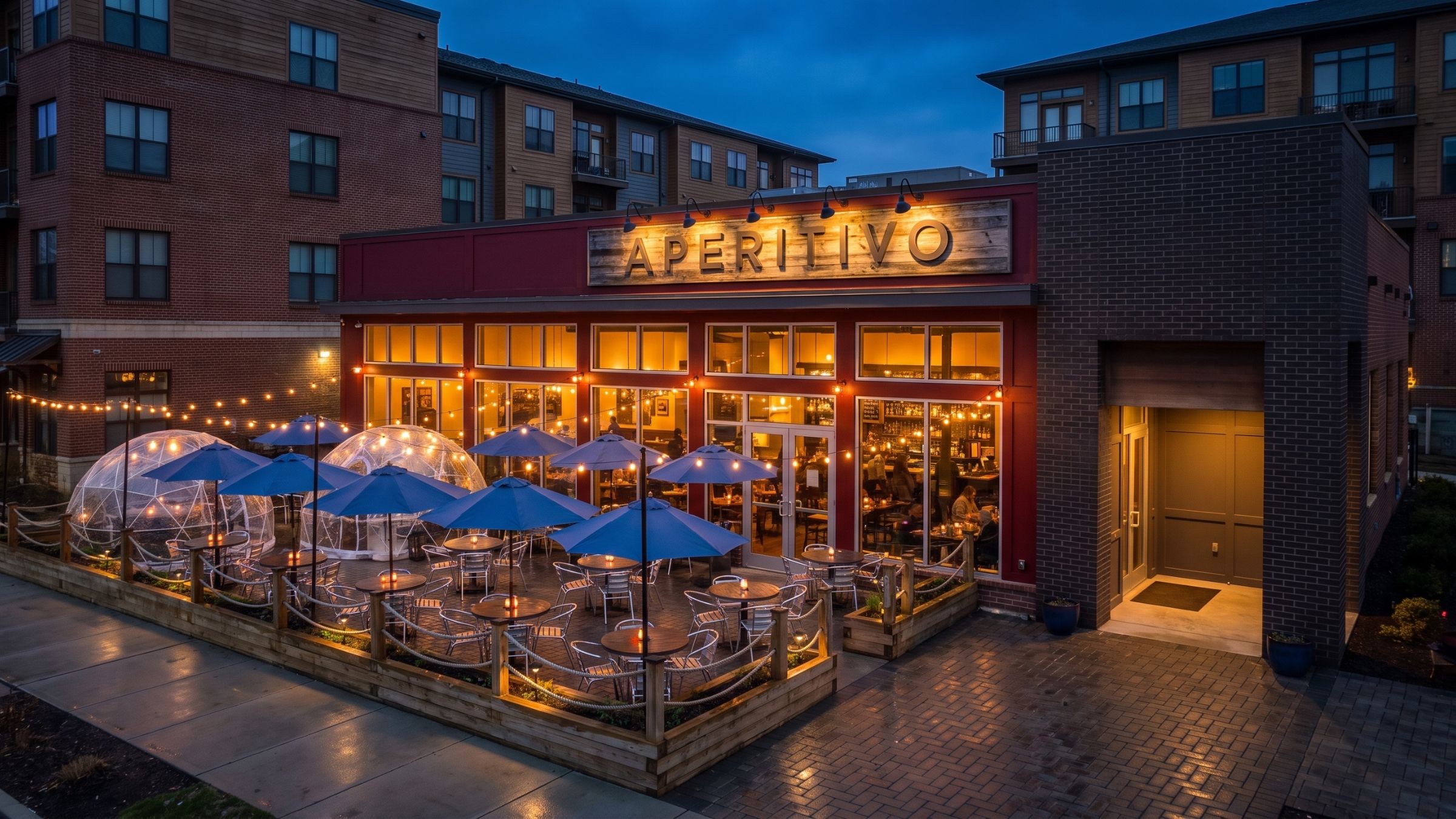 The Aperitivo patio at dusk, its windows and string lights glowing warm against a deep blue sky, with blue umbrellas and clear dome igloos on the paver terrace.