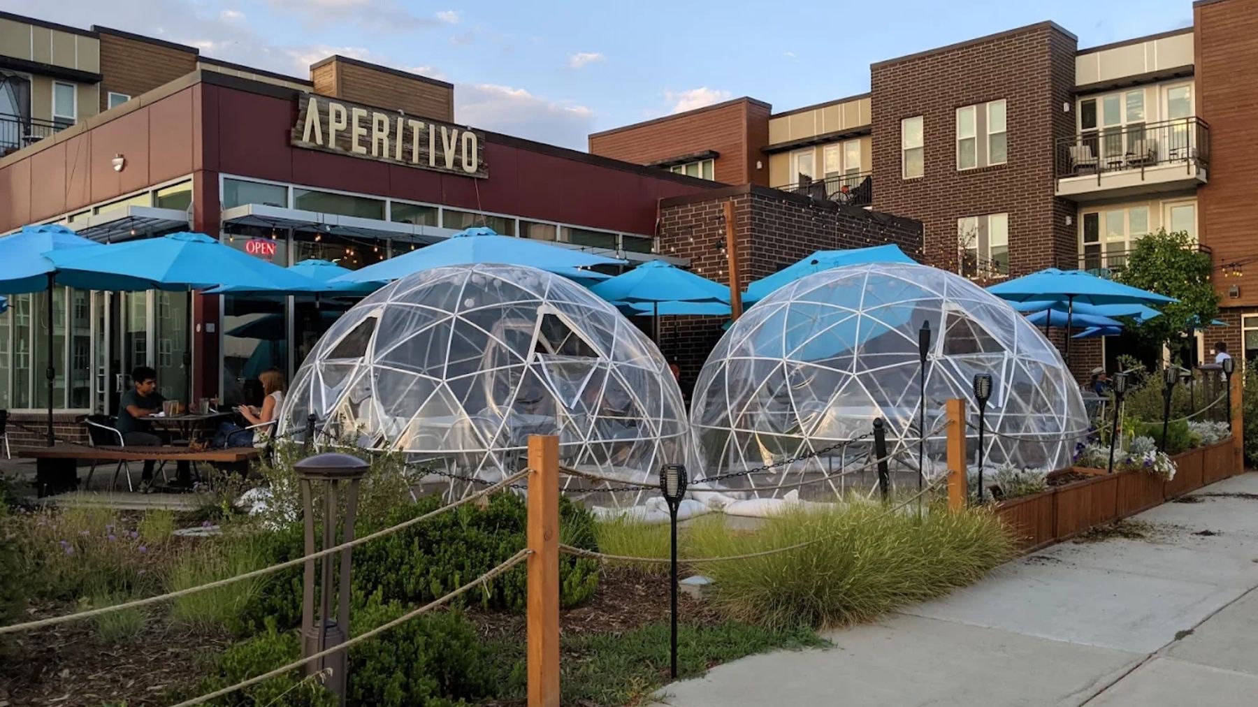 Two clear geodesic dome igloos set on the Aperitivo patio in the evening, with blue umbrellas and the lit sign behind them.
