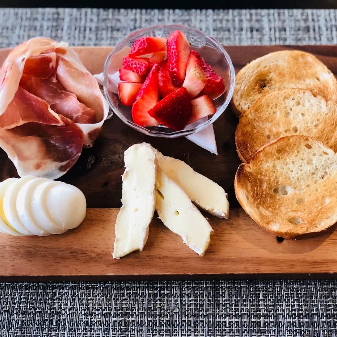 A wooden board laid with prosciutto, sliced soft cheeses, a glass of cut strawberries and rounds of toasted baguette.