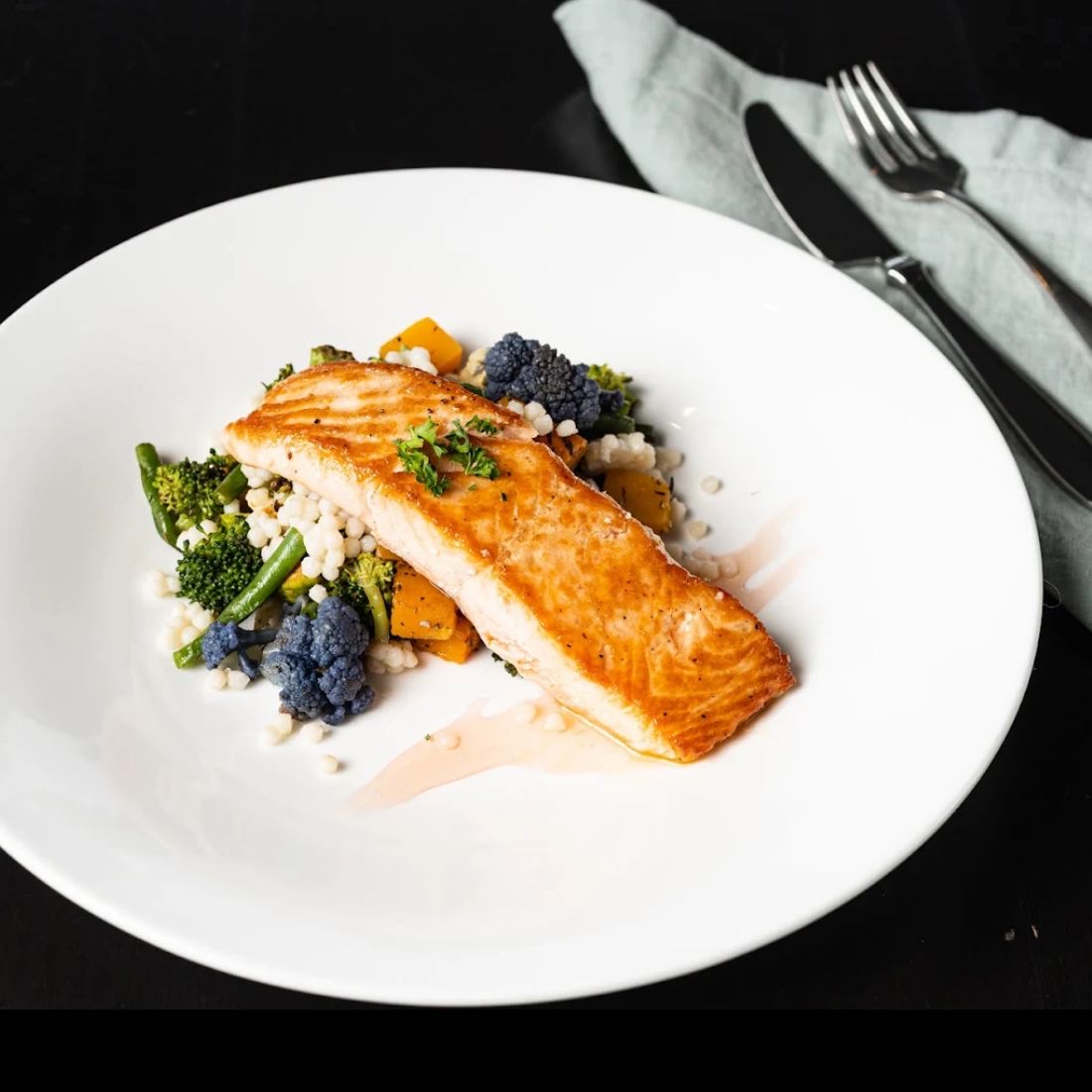 A seared salmon fillet resting on pearl couscous with roasted broccoli, purple cauliflower and squash, on a wide white bowl.